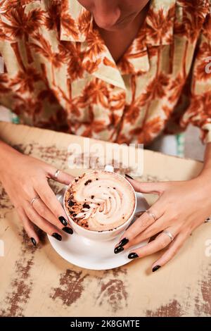 From above young female with black nails touching cup of fresh latte while sitting at cafeteria table Stock Photo