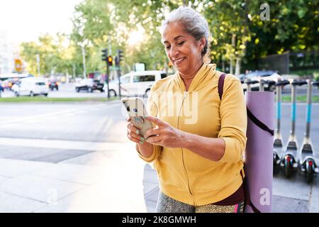 Positive elderly female athlete in sportswear with rolled up map smiling and browsing cellphone while standing near electric scooters after fitness tr Stock Photo