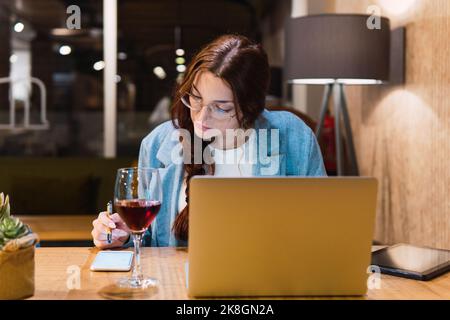 Smart young woman in glasses reading and analyzing data on smartphone while sitting at table with gadgets and red wine during work in restaurant in ev Stock Photo