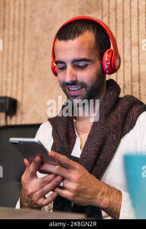 Happy man with wireless headphones smiling and text messaging on smartphone while working remotely from cafe Stock Photo