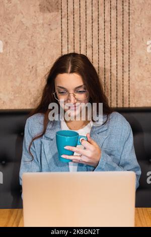 Positive young businesswoman with cup of coffee smiling and analyzing data on netbook during remote work from cafe Stock Photo