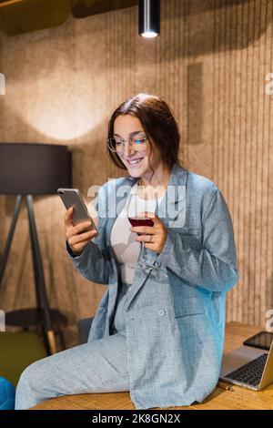 Young female manager in smart casual clothes with glass of red wine smiling and browsing social media on cellphone while sitting on table near laptop Stock Photo