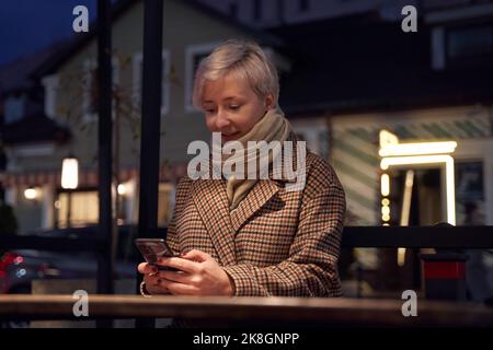 Young happy female in scarf browsing on smartphone while sitting in cafe near road with cars by night Stock Photo