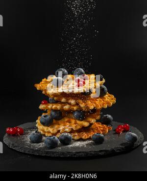 Fresh crispy Belgian waffles with fresh blueberries, blackberries, cranberries and red currants are sprinkled with powdered sugar on a black stone pla Stock Photo