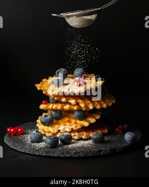 Fresh crispy Belgian waffles with fresh blueberries, blackberries, cranberries and red currants are sprinkled with powdered sugar on a black stone pla Stock Photo