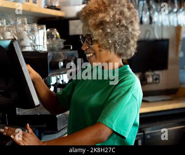 Happy African American female waitress using cash register while working in a pub. Stock Photo