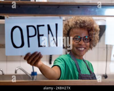 Happy African American woman looking at camera and Hanging Open Sign by the Glass Window Stock Photo