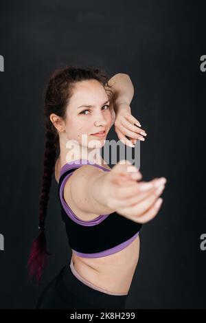 A pretty brunette girl dancing. Smooth body movements. Young sports woman stretches her arms towards the camera. Dark backgroud. Femininity concept. Stock Photo