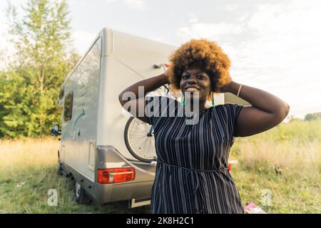 Body positive and independent Black woman looking at camera, holding her hands in her afro hairstyle hair, and standing in front of her white van. Outdoor shot. High quality photo Stock Photo