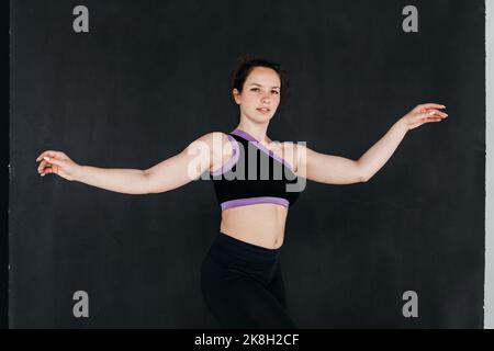 A pretty brunette girl dancing. Dark backgroud. Studio portrait of a young sports woman in a black pants and top. Femininity concept. Stock Photo