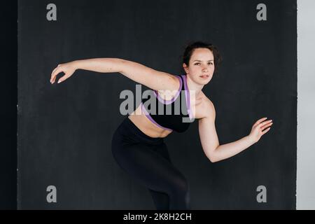 A pretty brunette girl dancing. Dark backgroud. Studio portrait of a young sports woman in a black pants and top. Femininity concept. Stock Photo