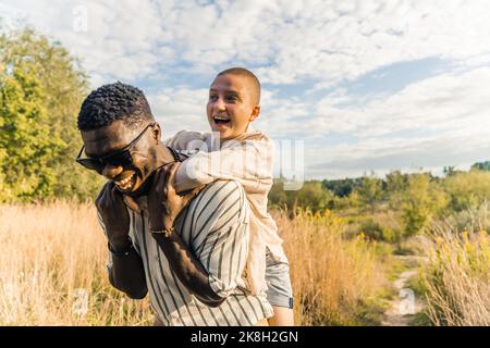 Piggyback surprise. Tall Black handsome man in sunglasses and white, stripped shirt having fun with his caucasian friend in the middle of country meadow. Beautiful memories from summer. High quality photo Stock Photo