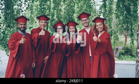 Portrait of excited graduating students multiethnic group standing outdoors in red gowns showing thumbs-up and looking at camera. Stock Photo