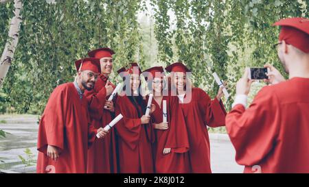Multiethnic group of graduating students is posing for smartphone camera, waving hands with diplomas and shouting while guy is shooting them touching screen and gesturing. Stock Photo