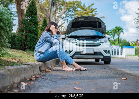 Asian woman sitting beside car after a car breakdown on street. Concept of vehicle engine problem or accident and emergency help from Professional mec Stock Photo