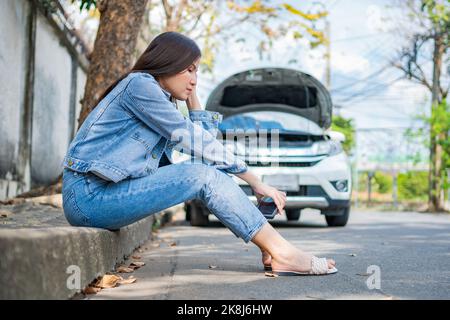 Asian woman sitting beside car after a car breakdown on street. Concept of vehicle engine problem or accident and emergency help from Professional mec Stock Photo