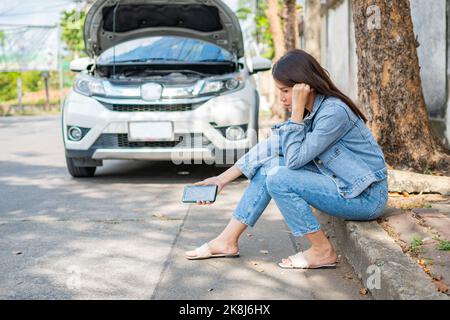 Asian woman sitting beside car after a car breakdown on street. Concept of vehicle engine problem or accident and emergency help from Professional mec Stock Photo