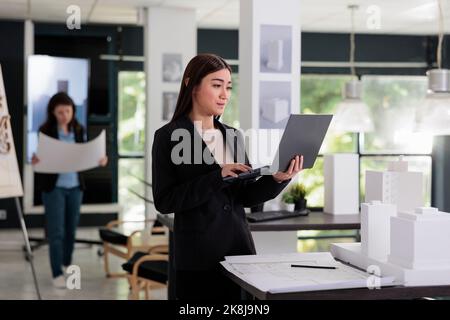 Architect modeling 3d building, using software on laptop, working with city construction project in architecture office. Employee engineering, real estate worker standing with computer Stock Photo