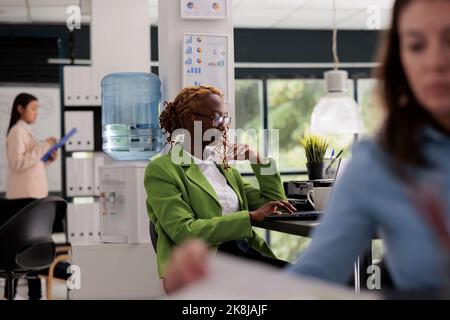 Thoughtful employee working business office open space, manager sitting at workplace desk, colleagues on background. Young woman thinking, typing on computer, side view medium shot Stock Photo