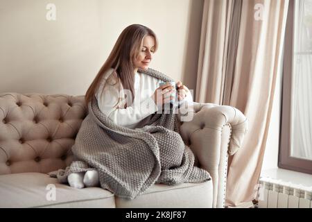 Woman sitting at home in beige minimalistic living room on sofa, drinking tea or coffee from blue mug, dressed in knitted plaid, Cozy hygge. Caucasian Stock Photo