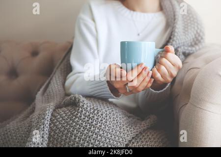 Close up of hands holding blue cup of tea or coffee. Model in white sweater and cozy plaid is sitting, relax at home on sofa. Depth of field, empty sp Stock Photo