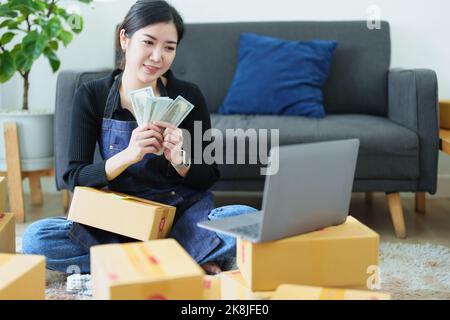 Starting small business entrepreneur of independent Asian woman smiling and holding money using laptop computer with cheerful success of online Stock Photo