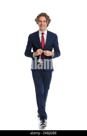 picture of happy young busienssman with curly. hair and blue eyes unbuttoning and opening suit while smiling and walking in front of white background, Stock Photo