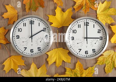 Two clocks, one shows two o'clock, the other shows three o'clock. Symbol of time change. Autumn fallen leaves. Stock Photo