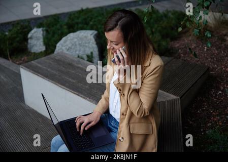 Woman remotely working, sitting on bench at city street and using laptop. Female freelancer work online, typing on laptop and talking with smartphone Stock Photo