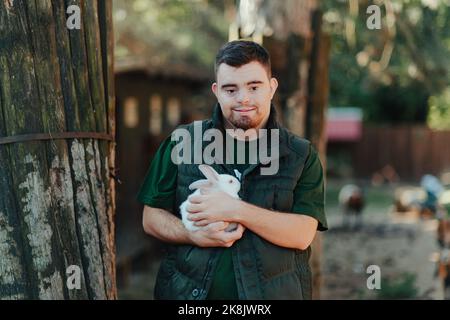 Caretaker with down syndrome taking care of animals in zoo, stroking rabbit. Concept of integration people with disabilities into society. Stock Photo