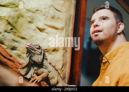 Caretaker with down syndrome taking care of animals in zoo, stroking iguana. Concept of integration people with disabilities into society. Stock Photo