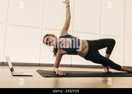 Smiling senior woman doing a side plank during an online yoga class. Elderly woman practicing a stretching asana on an exercise mat. Happy woman follo Stock Photo