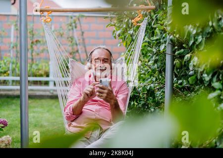 Happy senior man sitting in outdoor garden swing with headphones and scrolling smartphone. Stock Photo