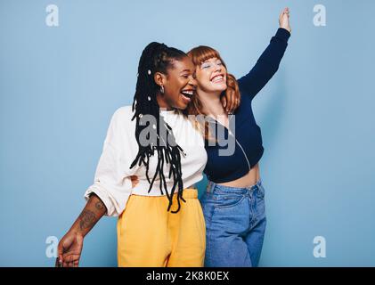 Two best friends laughing and having fun while embracing each other in a studio. Happy young women enjoying themselves while standing against a blue b Stock Photo