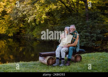 Senior couple in love sitting together on bench near lake, during autumn day. Stock Photo