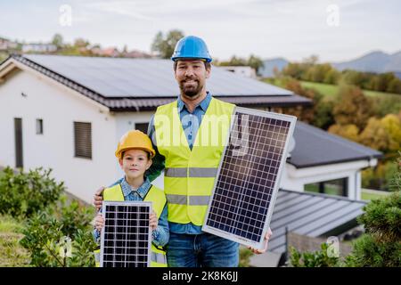 Smiling handyman solar installer standying with his daughter and holding solar module while installing solar panel system on house. Stock Photo