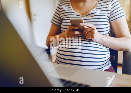 Young Caucasian pregnant woman using smart phone while sitting at table. On table laptop. Working pregnant women concept. Stock Photo