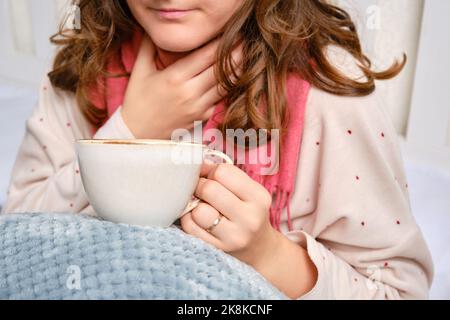 A sick woman is sitting on her home bed with a cup. Adult ill woman with a red scarf sitting on a bed in a white bedroom, female aged 35 years Stock Photo