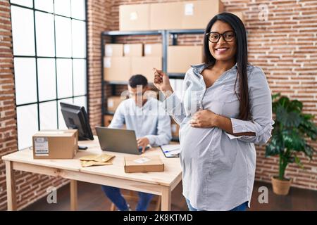 Young hispanic woman expecting a baby working at small business ecommerce smiling happy pointing with hand and finger to the side Stock Photo