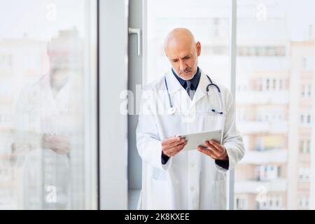 Portrait of careworn male doctor holding a digital tablet in his hand while standing on hospital’s foyer. Stock Photo