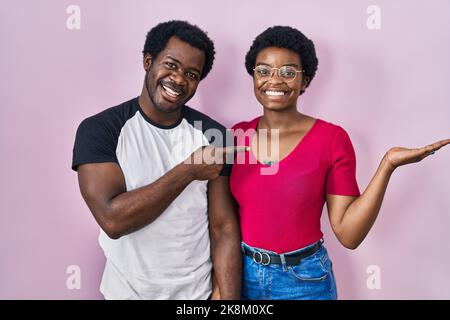 Young african american couple standing over pink background amazed and smiling to the camera while presenting with hand and pointing with finger. Stock Photo
