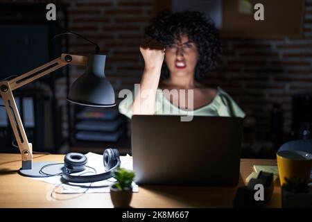 Young brunette woman with curly hair working at the office at night angry and mad raising fist frustrated and furious while shouting with anger. rage Stock Photo