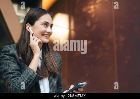 Charming business woman in headphones smiling looking away standing outside in front of the office building. Remote work, business, freelance, online Stock Photo