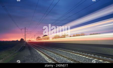 Modern railway at beautiful sunrise. Light trail of high speed train on railroad track. Moving modern intercity passenger train. Stock Photo