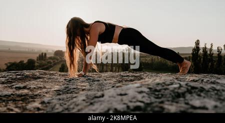 Well looking middle aged woman with long hair, fitness instructor in leggings and tops doing stretching and pilates on the rock near forest. Female Stock Photo