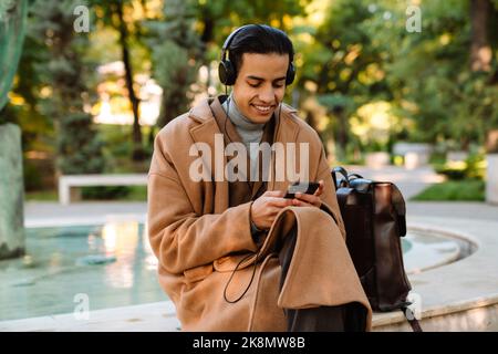 Young man in headphones smiling and using mobile phone while resting in park Stock Photo