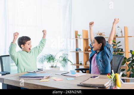excited kids showing win gesture near notebooks and paper notes on table at home,stock image Stock Photo