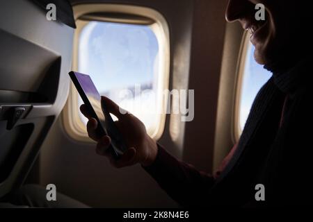 Businessman sitting next to airplane window and using smartphone. Depersonalized man fly on plane and reading with phone in online app. Person using Stock Photo