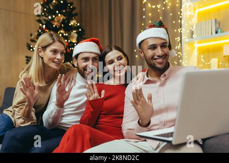 diverse friends celebrating New Year and Christmas together home, Christmas party with guests, men and women smiling and happy together talking on video call greeting friends remotely, using laptop. Stock Photo