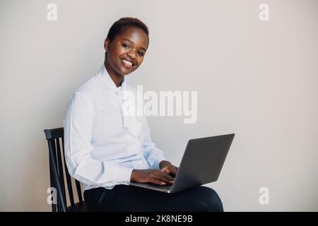 Cheerful African-american woman using laptop, having online meeting, sitting on chair against white wall. Looking and smiling at camera. Laughing Stock Photo
